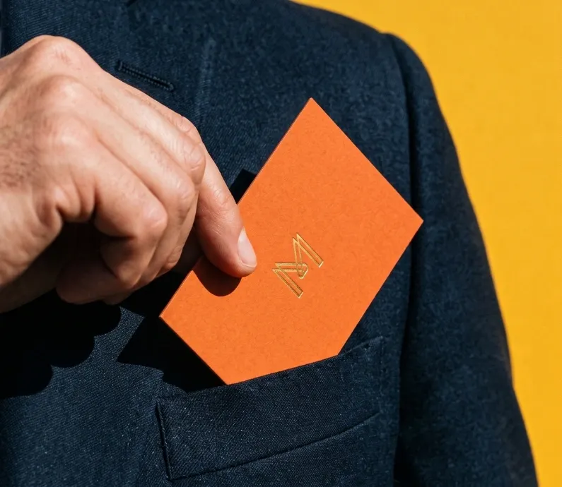 A hand sliding an orange business card with a gold foil mark into a navy jacket pocket